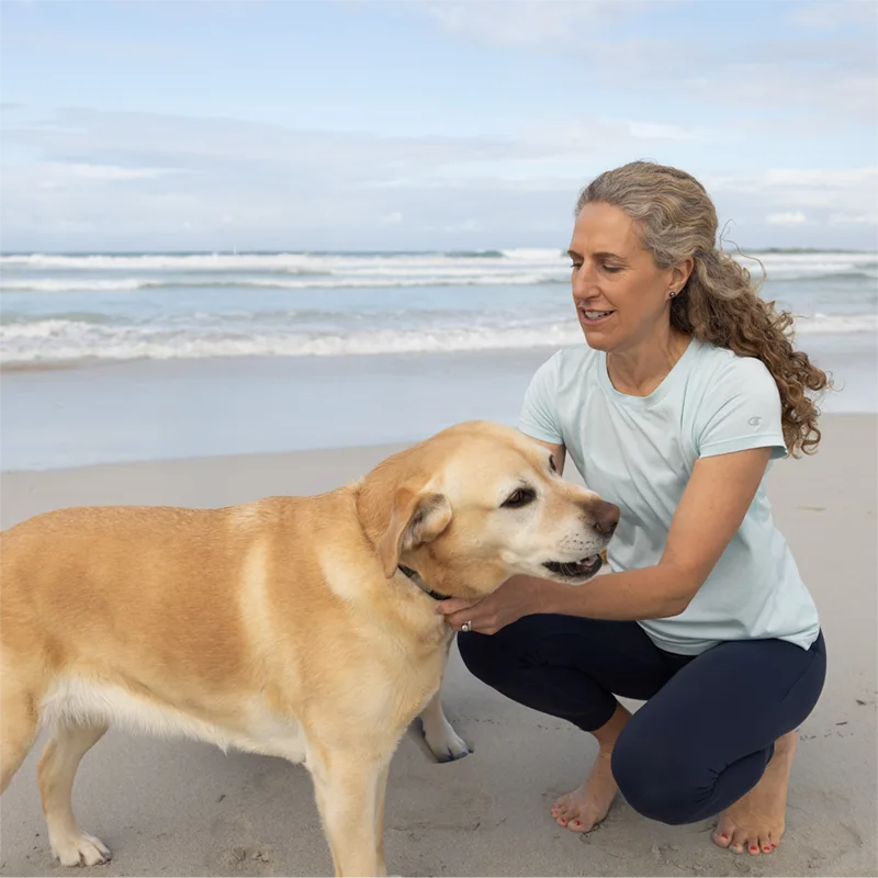 Lady crouching down near golden retriever dog on beach. Sally McKenzie, Pelvic Health Physio